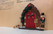 A miniature red door decorated with a little tartan bow and garland with a little wooden guard standing beside it.